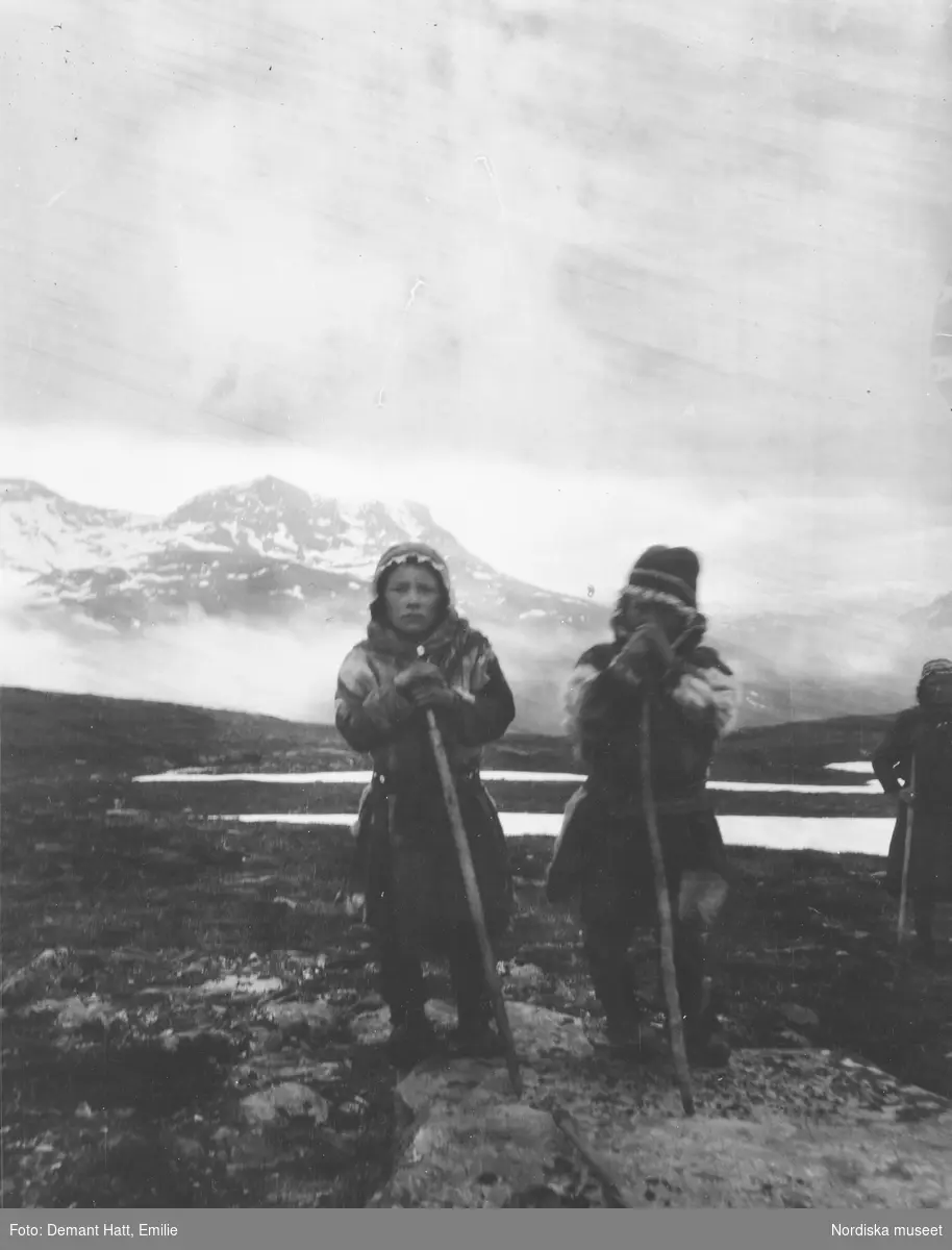 Tre barn/ungdomar med vandringsstavar ute i naturen. I bakgrunden fjäll. Vårflyttningen från Närvä. Bilden ingår i en serie fotografier tagna av Emilie Demant Hatt i Sapmi mellan åren 1907 och 1916.