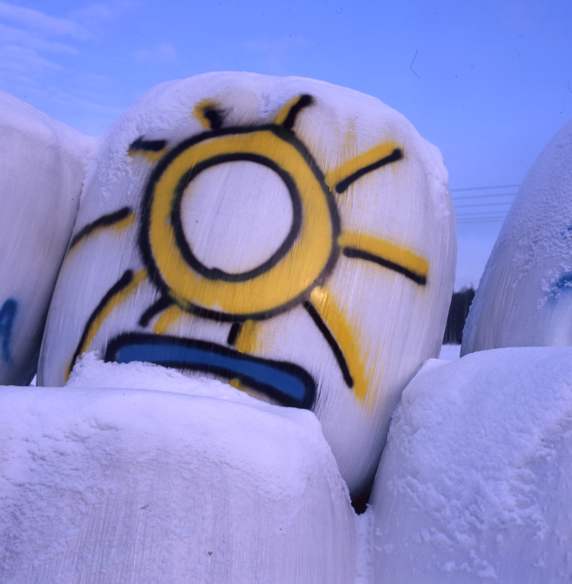 Inplastade ensilagebalar täckta med snö. På en av dom har någon ritat en stor sol.