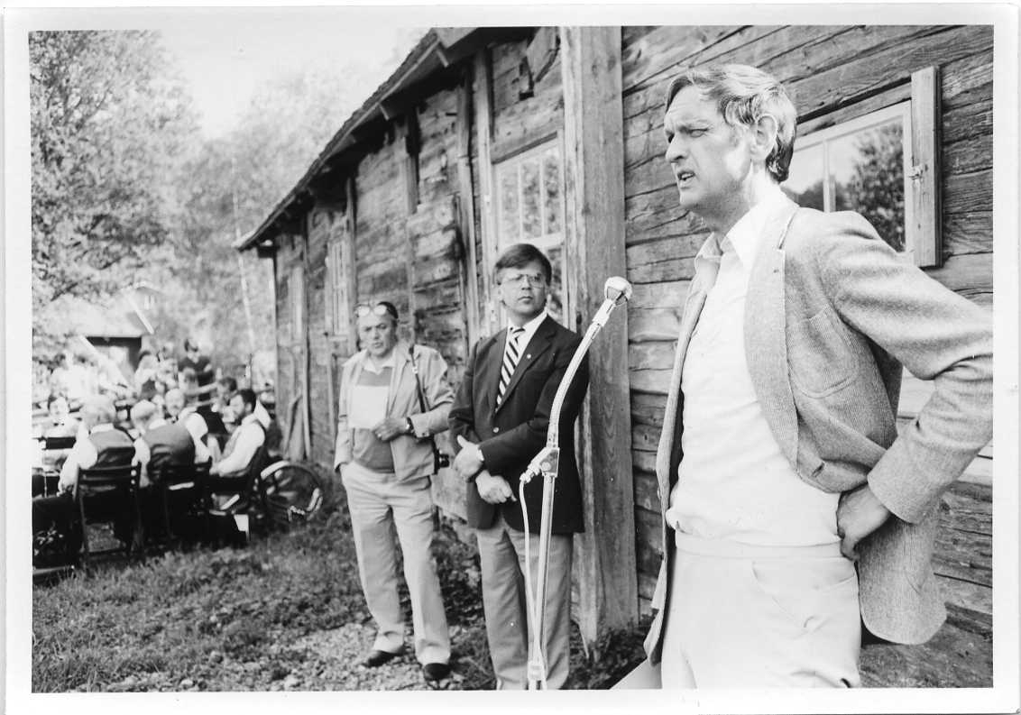 Tre män står utmed ladugården Rasten i Röttle. De är museichefen Olof Fong, museistyrelsens ordförande samt Rolf von Otter som talar i en mikrofon på stativ. I bakgrunden sitter blåsorkestern Juneblecket.