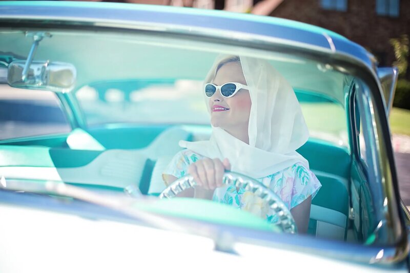 Bilde av dame med sjal på hode og solbriller i en åpen veteranbil