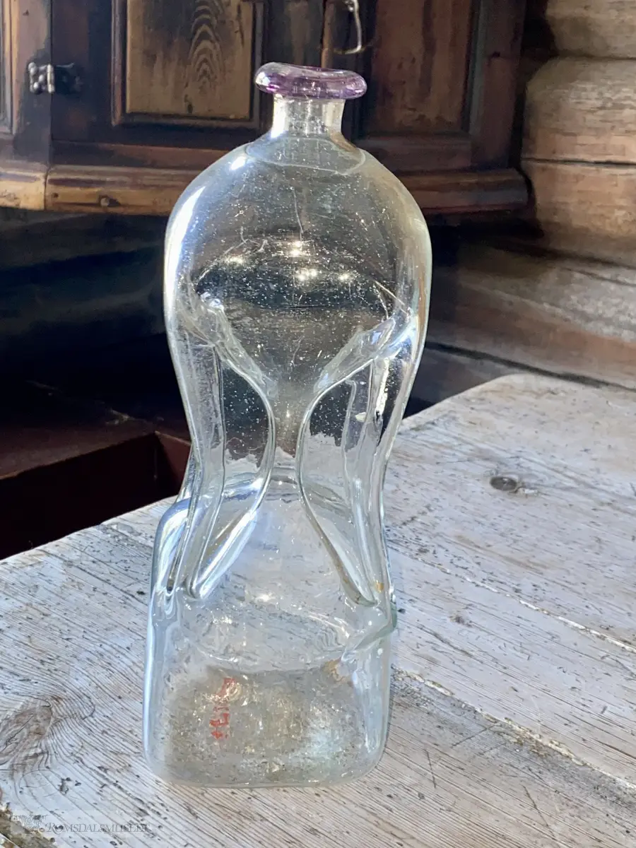 Håndblåst, timeglassformet glassflaske med puntelmerke. Glassplater mellom rørene som binder sammen øvre og nedre del.