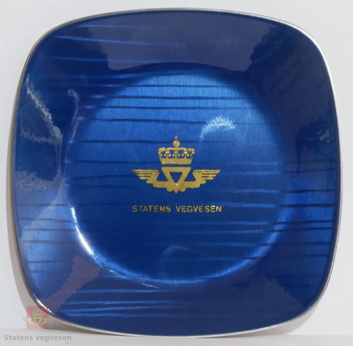 Skålformet lite blått askebeger i metall med Statens vegvesens logo i gull. På undersiden er det ubehandlet metall. Firkantet utførelse med avrundede hjørner.
