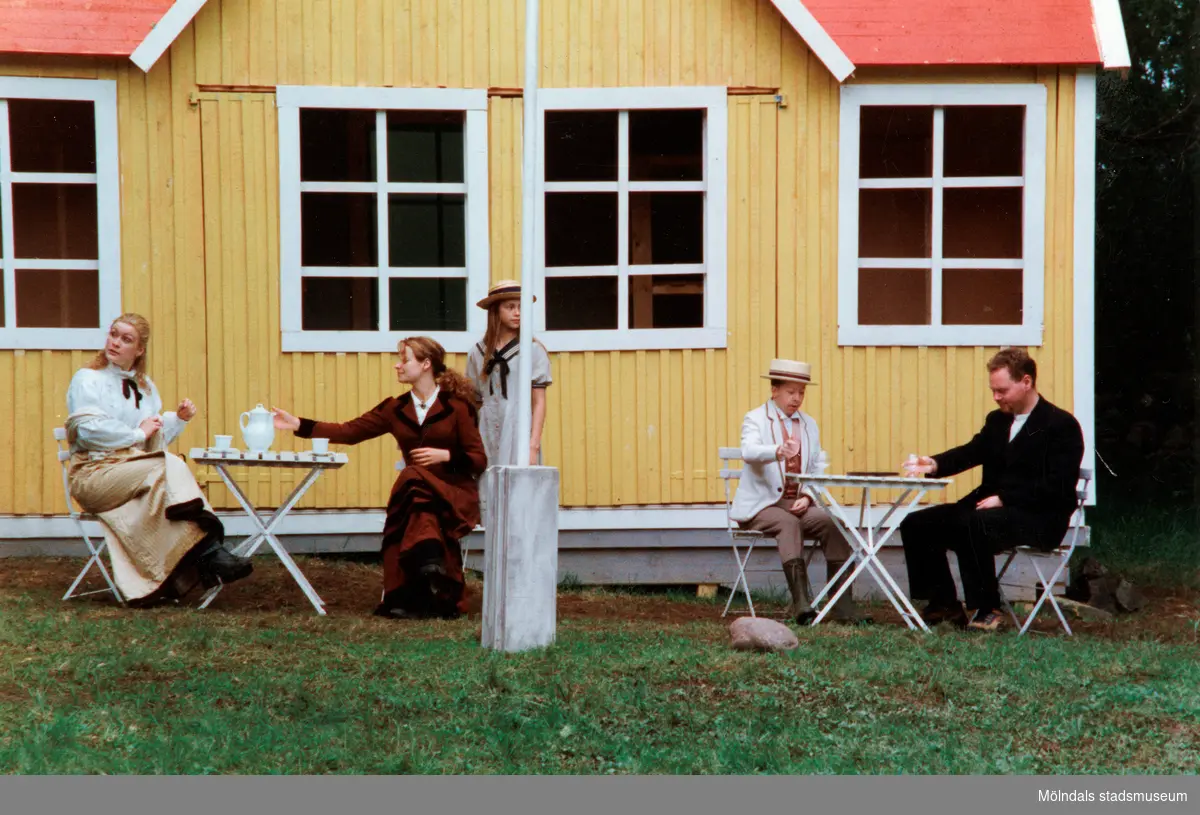 Sommarspelet/barnteaterföreställningen "Rasmus på luffen" 1999 vid Byxorna i Gunneboskogen (korsningen Christina Halls väg/John Halls väg). Två fikabord är uppdukade med koppar och fat framför ett gult litet hus. Vid vänstra bordet sitter två kvinnor, vid det högra sitter två män. I mitten står en flicka vid en flaggstång.