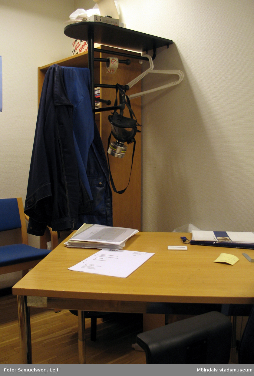 Ett rum med ett bord, en hylla samt ett par stolar. Från väggen hänger arbetskläder samt en skyddsmask. Interiörfotografi från byggnad vid Soabs industrianläggning i Mölndals Kvarnby, år 2007. Anläggningen användes vid fototillfället av Hexion Speciality Chemicals Sweden AB.