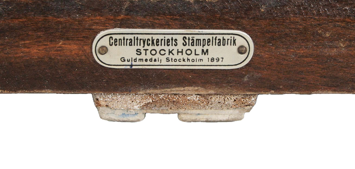 Gummistämpel "LRA", på kvadratisk brunbetsad träplatta. Avlångt, profilerat handtag. Framtill, påspikad oval, grå plåtskylt märkt: "Centraltryckeriets Stämpelfabrik STOCKHOLM,  Guldmedalj Stockholm 1897".
