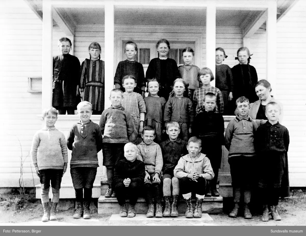 Gruppfoto med barn på brokvisten till ett ljusmålat hus, sannolikt ett skolfoto. Till höger en kvinna/lärarinna. Samma brokvist och kvinna som SuM-foto035238.