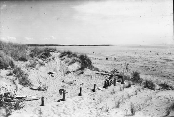 Kolorerat vykort, "Haverdalsstrand". Strandvy med sanddyna och badande i havet. I förgrunden en luftmadrass och barnvagn, på stranden några cyklar.