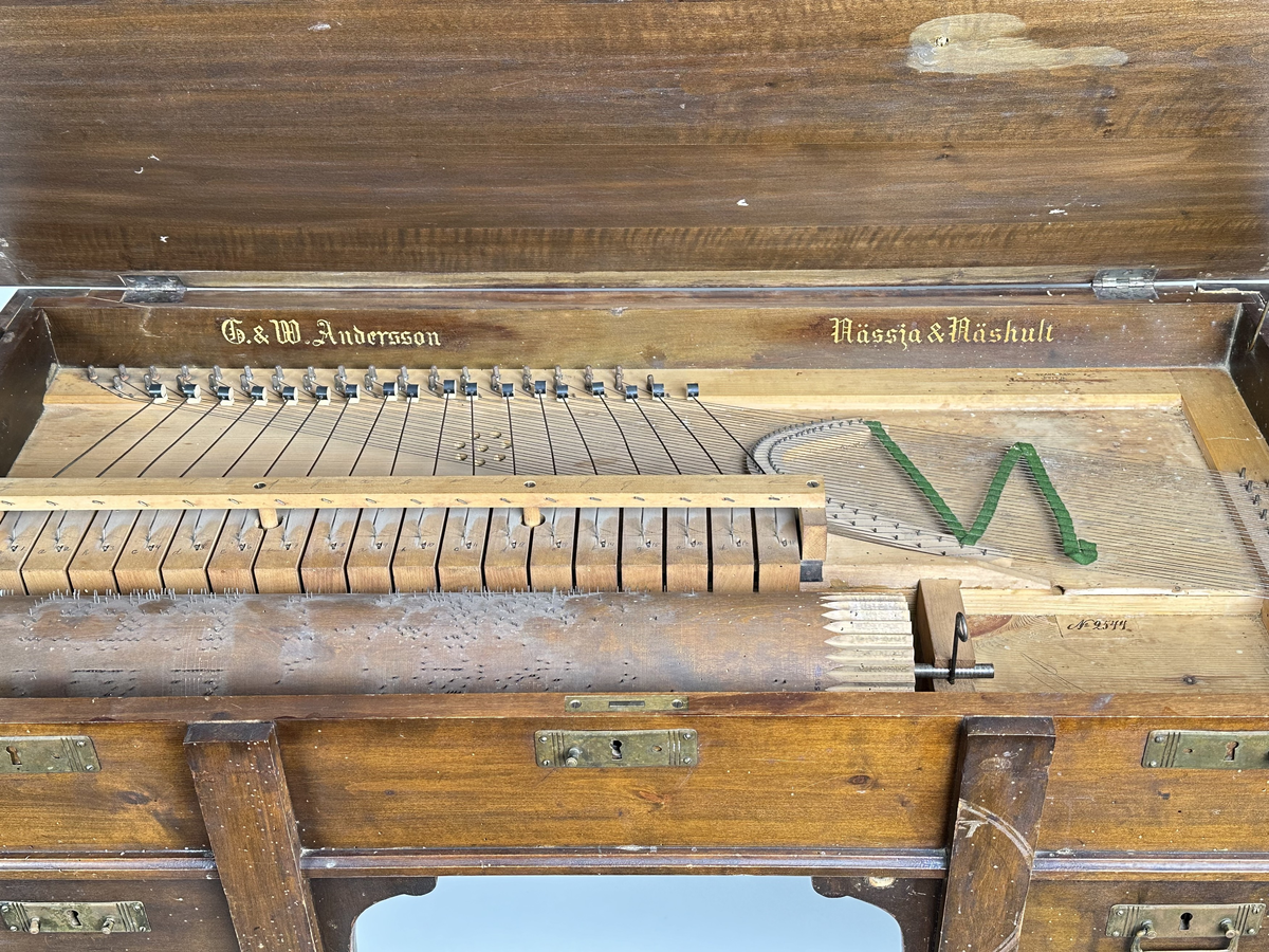 A) Pianoharpa, i form av ett skrivbord av mazarin-typ. Av mörkt betsat och polerat trä. Skrivbordet kraftigt angripet av trägnagare. Svarvade ben och hög sarg, med två utdragbara lådor och tre blindlådor - alla med mässingsbeslag. Bordsskivan uppfällbar och ställbar med metallhake, varunder spelverket med strängar och piggförsedd vals gömmer sig. 
B) och C), två valsar med olika melodier på. Cylinderformade, av trä, med mittaxel och piggar av metall. Valsen snurras med en vev, som lämnat ett svängt märke på möbelns front. 
D), vev av tunn metallarm med ett S-formad avslut i 45 graders vinkel från armen samt svarvad, balusterformat handtag av trä.
E) och F), stämnycklar av metall med fyrkantiga hål, samt enkla handtag av trä. 
G), sprint av metall för att säkra valsen på plats. Tunn, med ögla till avslut.
H), förvaringslåda för melodivals. Av trä, med inbyggd vagga för att hålla valsens mittaxel.