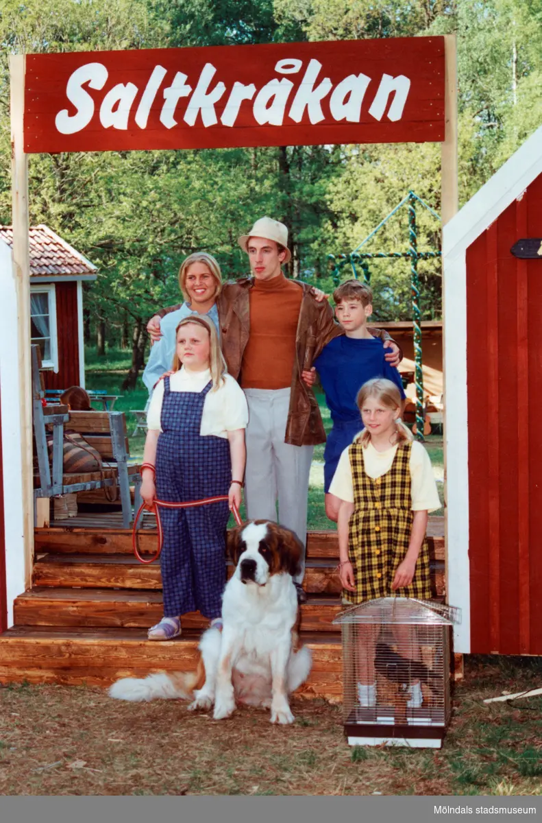 Sommarspelen/barnteaterföreställningen "Tjorven på Saltkråkan" 1996 vid Byxorna i Gunneboskogen. Två vuxna och tre barn (ett är Tjorven) samt hunden "Båtsman" poserar under skylten "Saltkråkan". En av flickorna håller en fågelbur med en korp i. Pappan i familjen spelas av Per Andersson. Relaterade motiv: 2024_0525 - 0533.