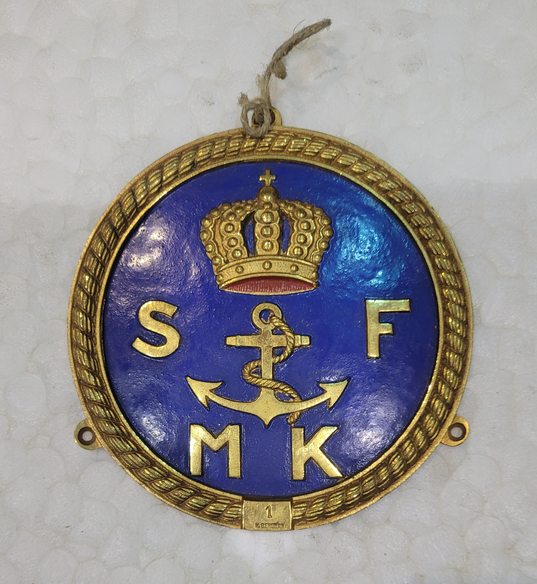 Stäv- eller skorstensmärke. Runt, gulmetall. Krönt ankare omgivet av bokstäverna SFMK mot blå botten innom krans. Nedtill stämplad I.H. Bergman.