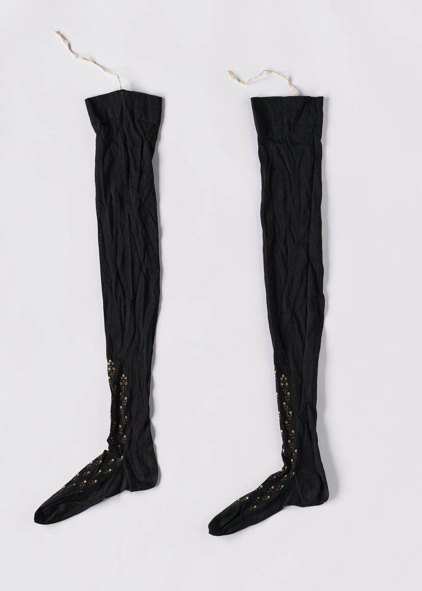 Ett par strikkede svarte nylonstrømper. Foten er formsydd. Det er broderinger i blomstermønster fra oversiden av foten og oppover leggen. Det er bånd i kanten, og hull i brettekanten for å tre inn bånd.