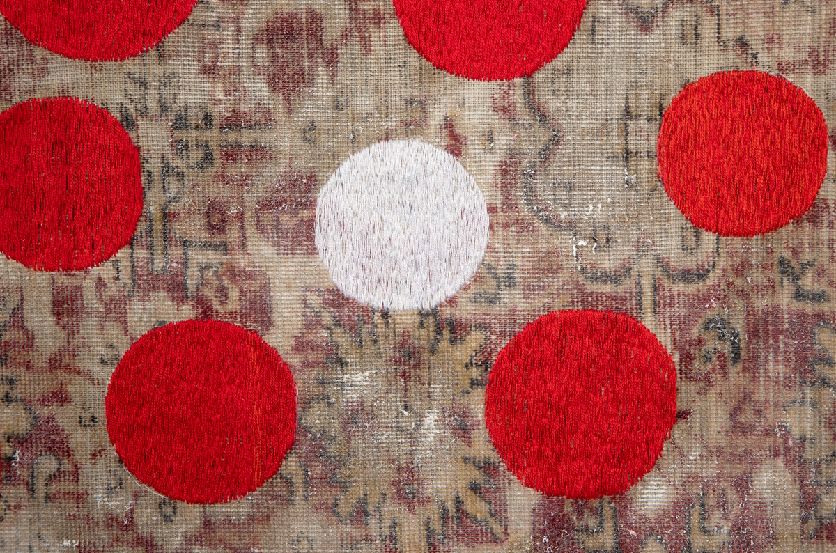 Kvadratisk veggtekstil av fløyel med trykt mønster som minner om persisk teppe. Stoffet har et tynt lag med hvit akrylmaling. Håndbroderte sirkler i hvit og rød bomullstråd.