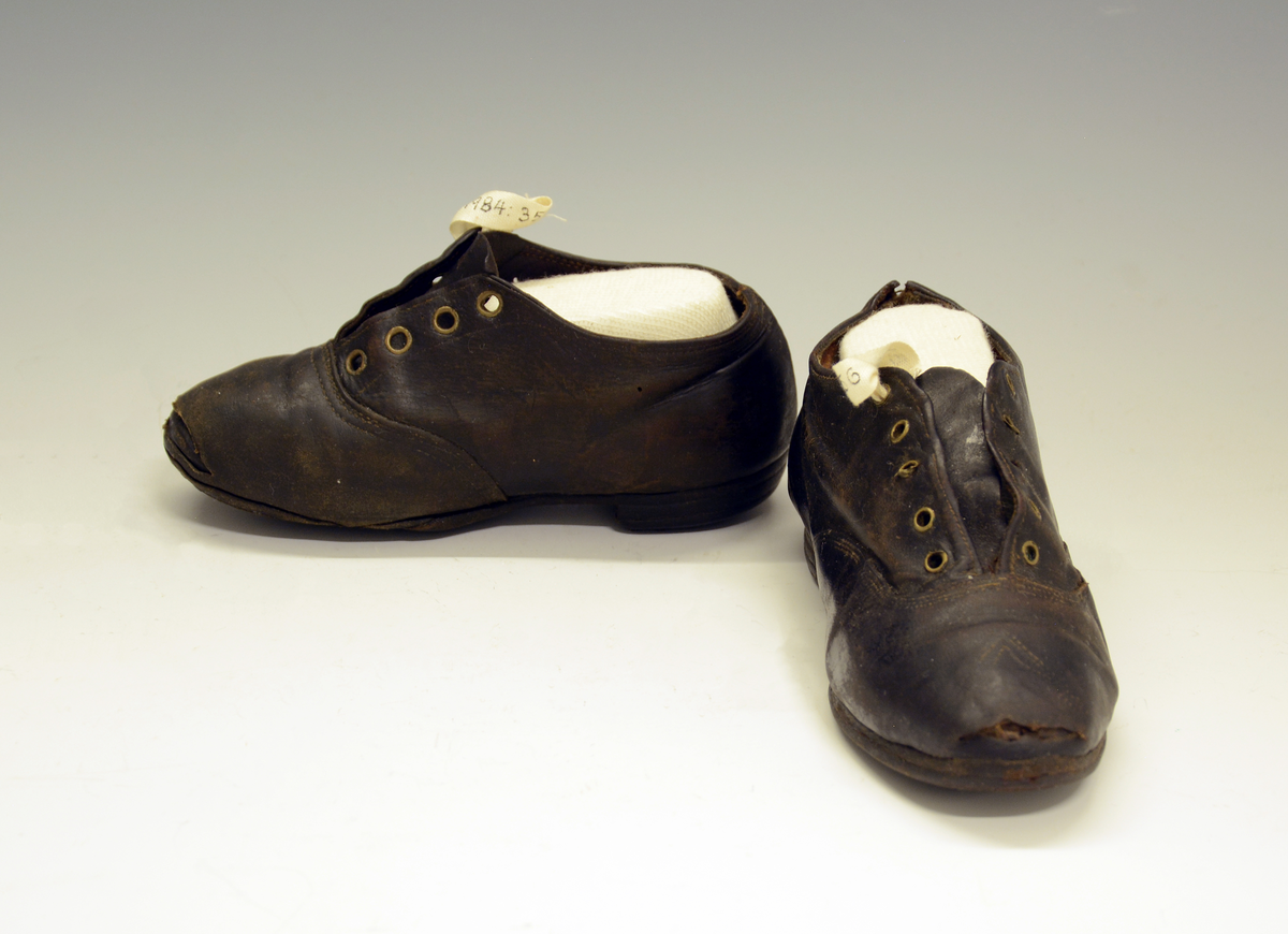 Barnesko. 1 stk sko i brunt lær med 4 stk snørehull med metallmaljer. Jernstifter i hæl og treplugger i sålen.