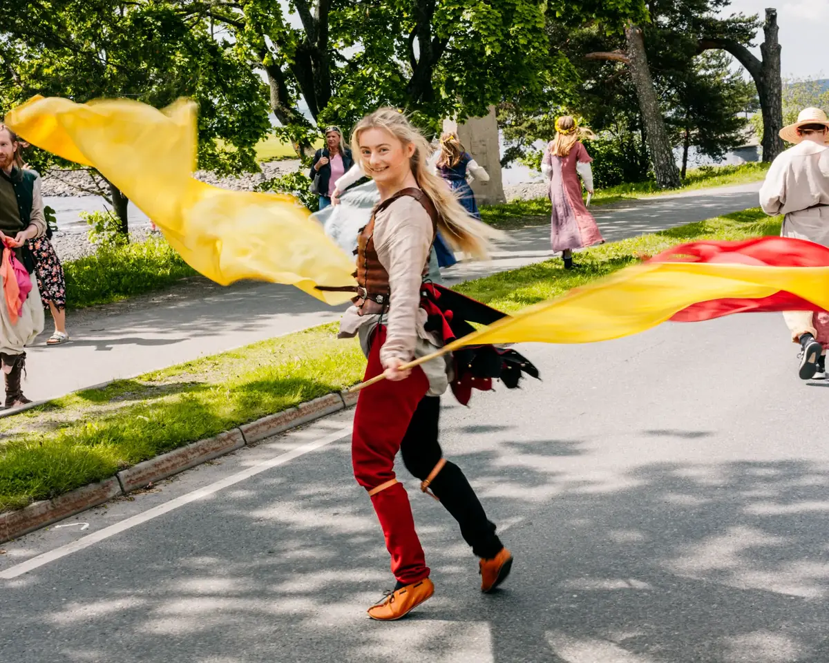 Ung kvinne med rødt, brunt, hvitt og svart middelalderkostyme som danser med røde og gule flagg i parade.