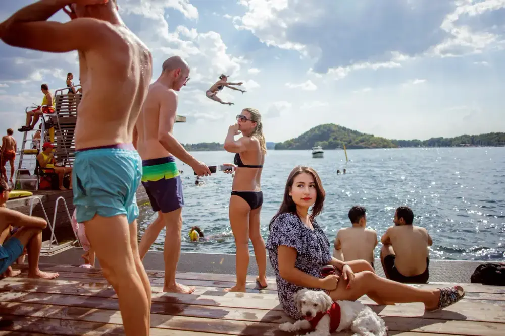 Bilde av mange mennesker delvis på en brygge og i vannet. En kvinne sitter i midten med en liten hvit hund ved sin side.