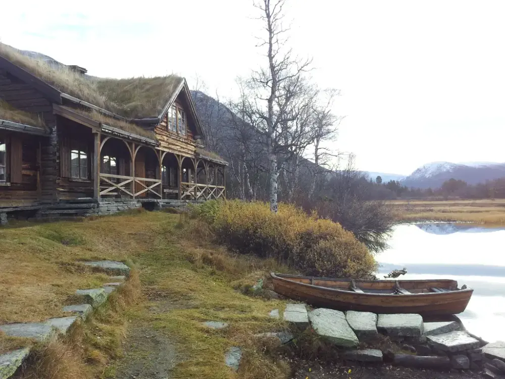 Tømmerhytte med svalgang ved fjellvann, en liten trebåt ligger på stranda.