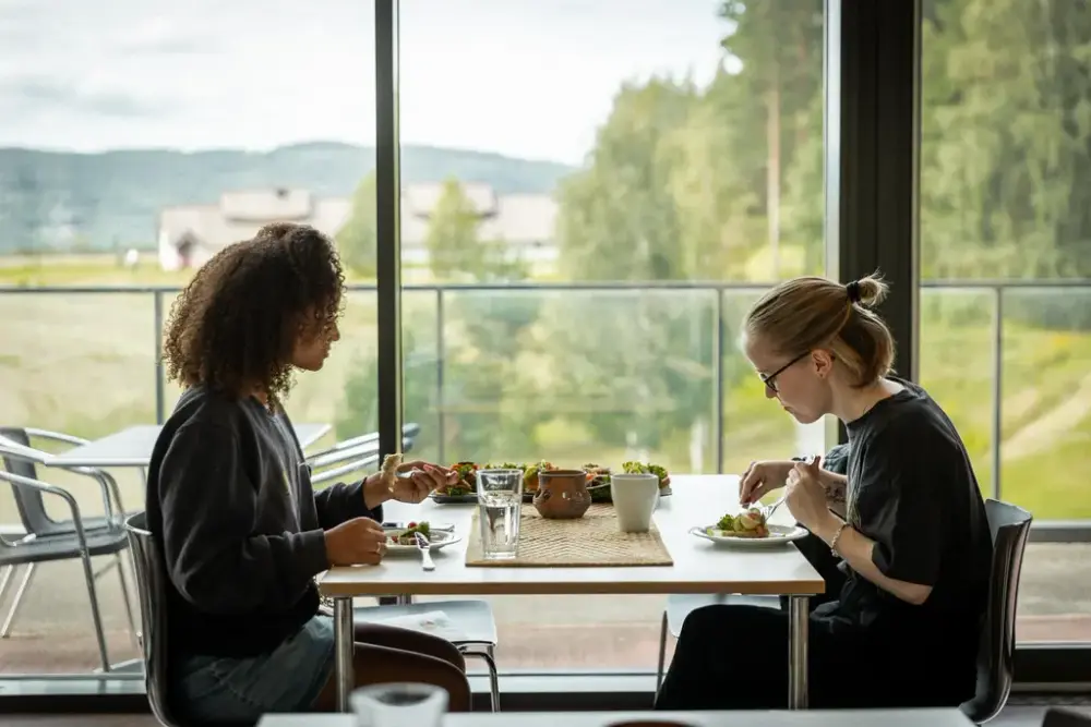 To damer sitter på museet og spiser lunsj. Utsikt mot langhuset 