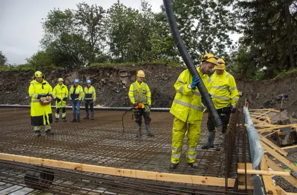 Sju arbeidere i signalklær og hjelm står på en støpt flate belagt med armeringsjern, to av dem styrer en slagne som spyr ut mer betong.