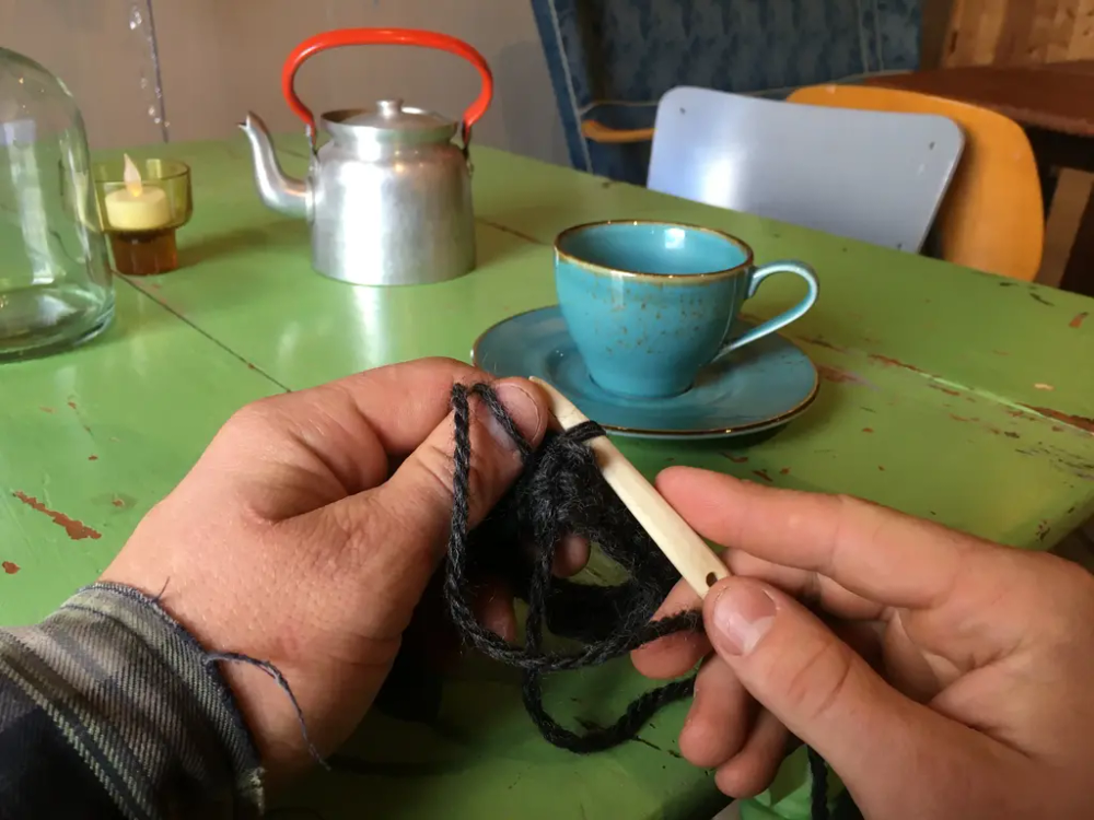 Hendene til en mann i forkant av bildet. Han holder på med nålebinding. I bakkant et grønt bord, kaffekopp og kaffekjele