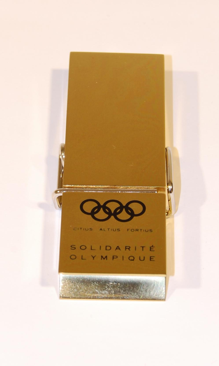 Gullfarget klype av metall med motiv av de olympiske ringene.
Det følger med en eske til klypen. 