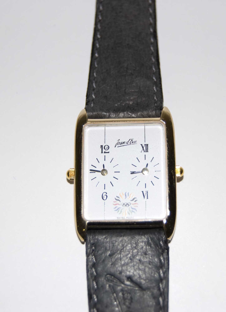 Gull- og sølvfarget armbåndsur med sort reim og hvit urskive. På urskiven er det et emblem med motiv av de olympiske ringene og en stilisert sol. Det følger med et grått etui.