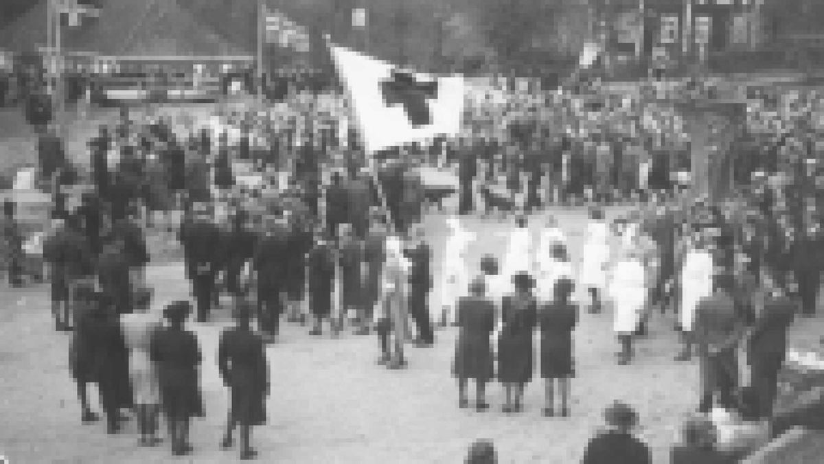 Eydehavn torv, 17. mai 1945
Vi ser murgårdene, norske flagg, Røde kors flagg og hvitkledd personell og mye folk  
