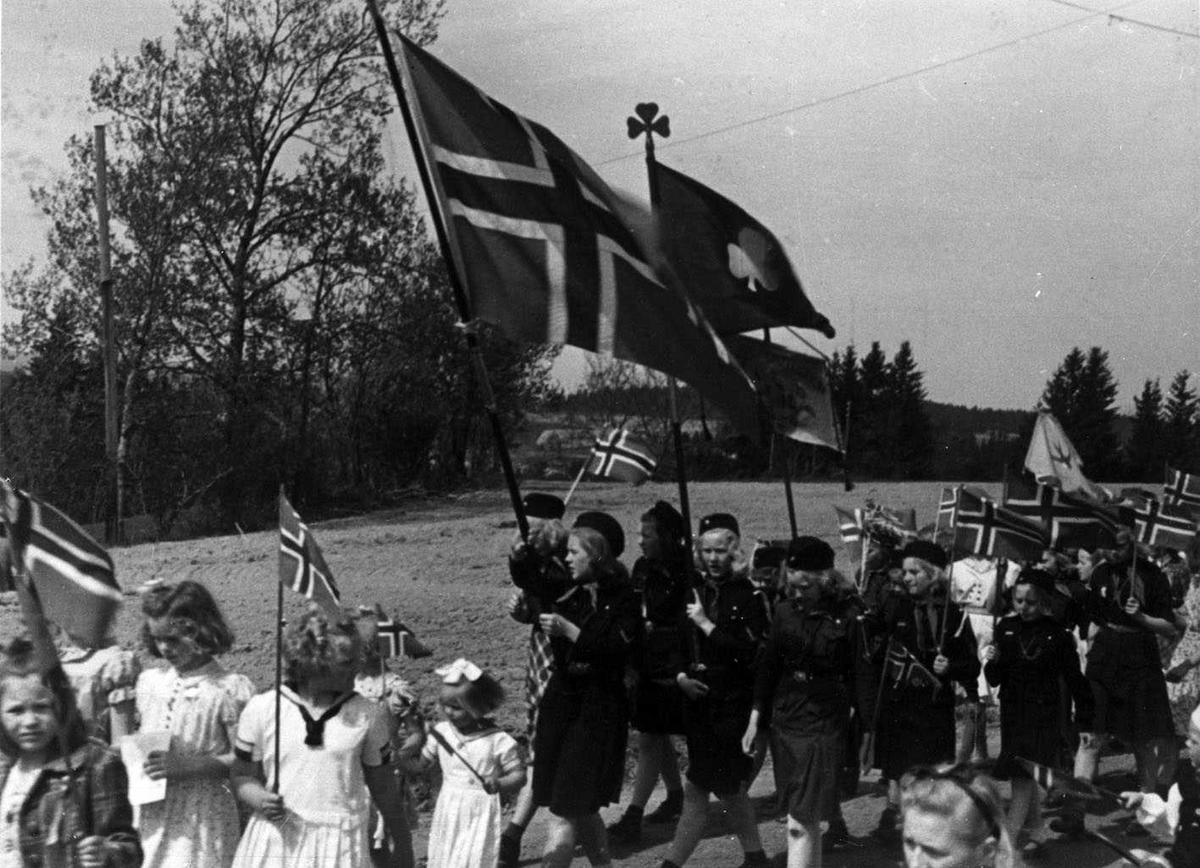 17. mai feiring ved Skedsmo kirke 1945. 17.maitog på marsj. Speidere i tog, fører kløverbanner i tillegg til norsk flagg.