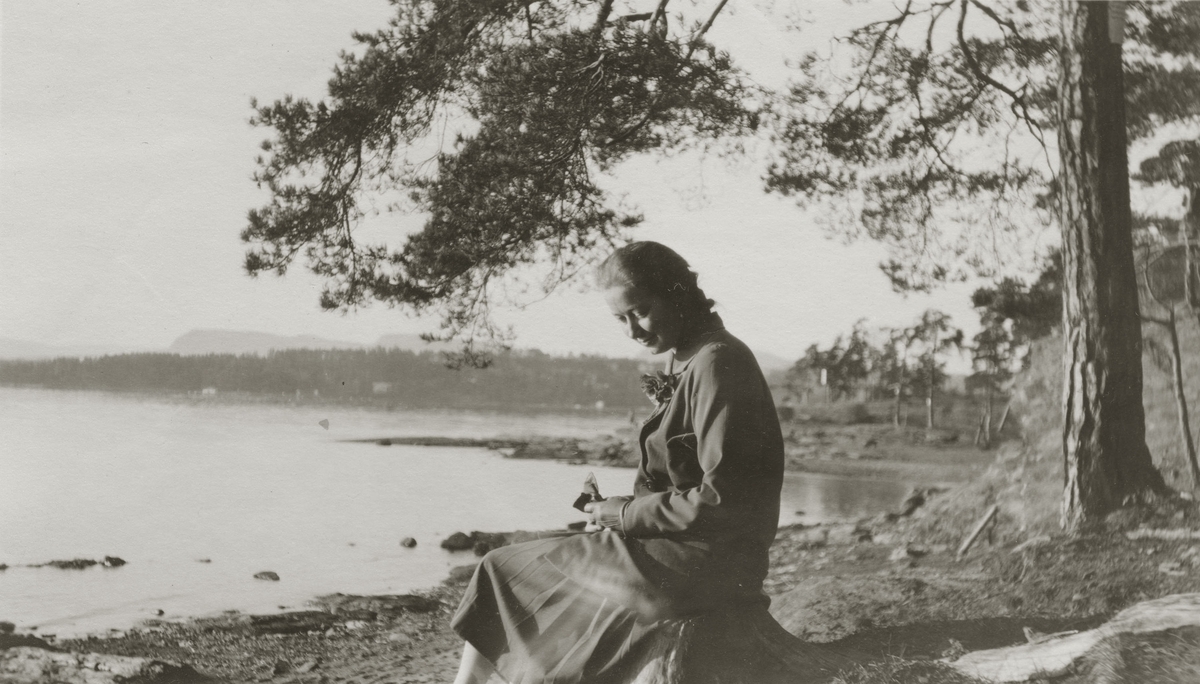 Antatt: Margit Frogner sitter på stubbe ved vannkanten. Skjørt, bleser, veske, hansker og blomst i jakkeslaget.