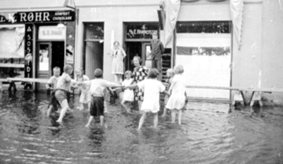 Strandgata, Hamar. Mjøsflom 1927. Barn ringlek i vannet, utenfor G. K. Røhnes og E. Hammerstads forretninger. Til h. på trappa Dagny Brænd, Ole J. Lie.