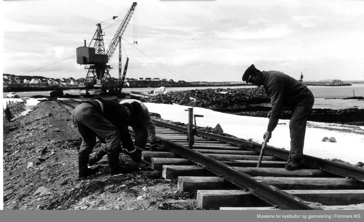 Åsmund Olsen, Oskar Olsen og Hans E. Hansen justerer jernbanen på Revnes. Ved hjelp av domkraft, hakker og spader fyller de under svillene, 1966