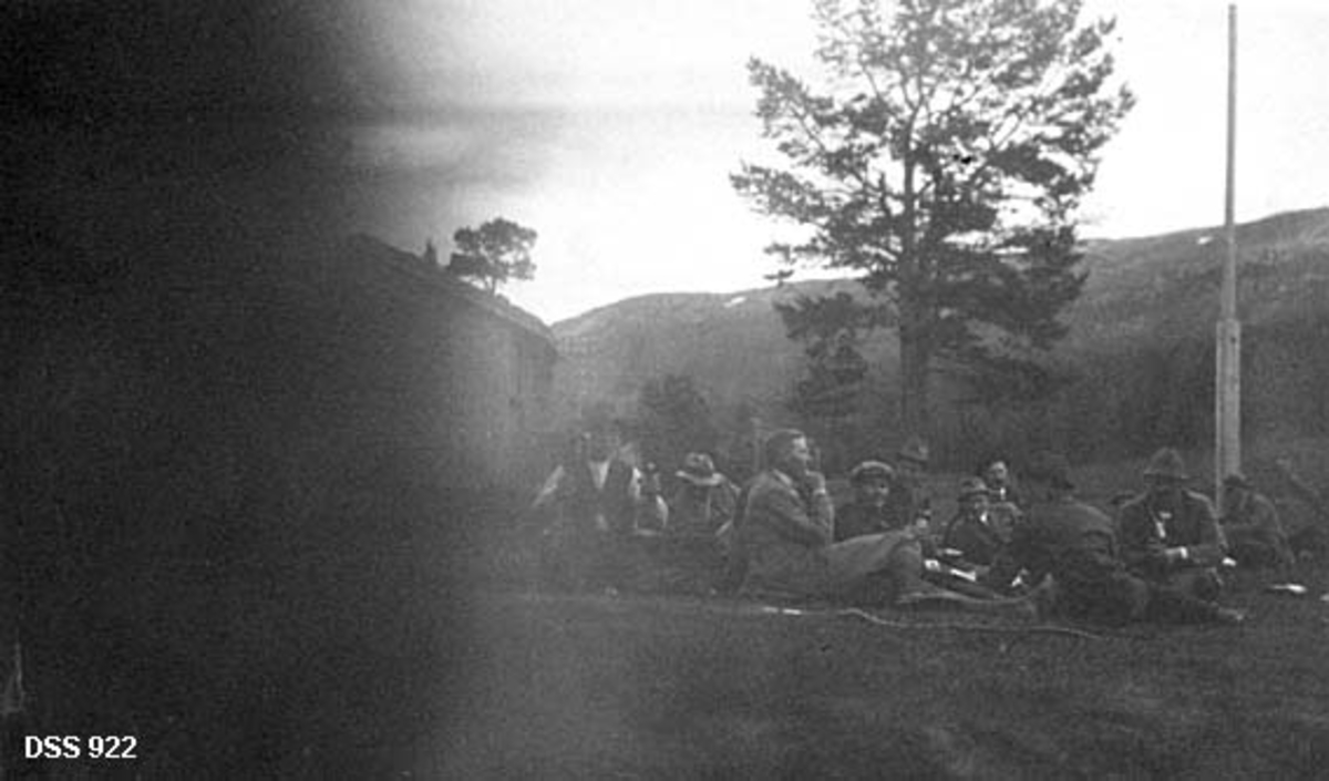 Rast i forbindelse med ekskursjon i Saltdal statsskoger sommeren 1919.  Fotografiet er tatt på en grasbakke ved Berghulnes.  Cirka 15 menn sitter på bakken og spiser og drikker.  I bakgrunnen ei flaggstang, ei stor furu og en husgavl.  Den venstre delen av bildeflata er fullstendig svart. 