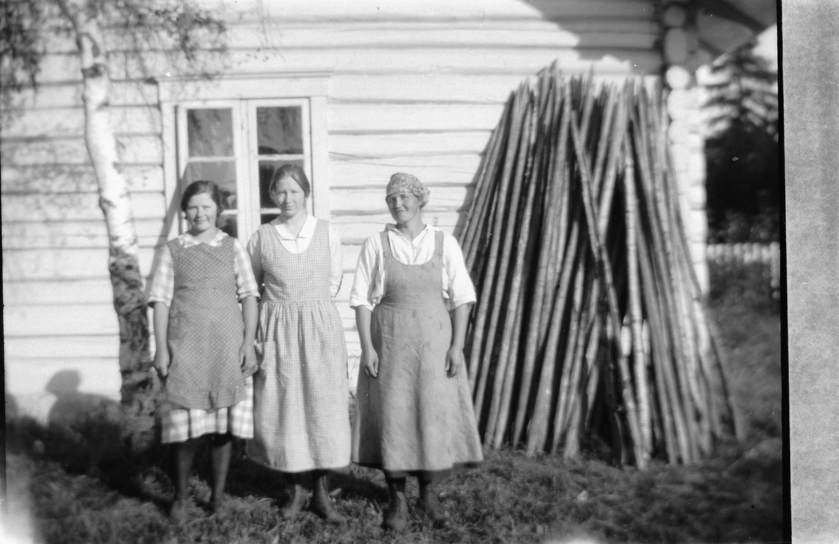 Tre kvinner ved en husvegg og en stabel med staur. 