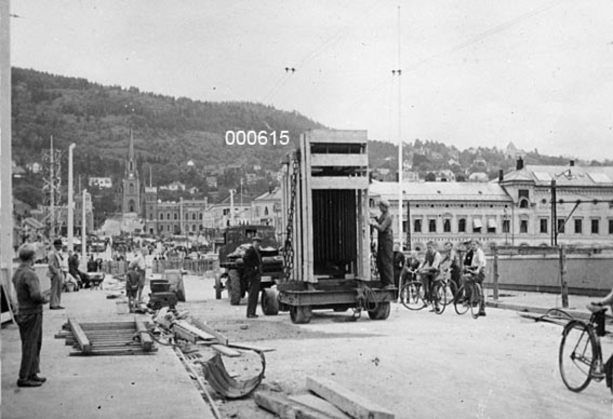 Transport av transformator som skulle installeres på A/S Buskerud Papirfabrikk på Åssida i Drammen (den gang i Lier kommune) i 1936.  Fotografiet er tatt på det som den gangen var ei ny bru over Drammenselva.  Transformatoren skulle åpenbart fraktes fra jernbanestasjonen over elva og videre på vegen mot Krokstadelva til en nådde fabrikken.  Ifølge et avisoppslag (jfr. SJF-F. 000618) var dette en transport som først ble mulig da det kom ny bru, fordi den gamle ikke var konstruert for å bære slike tunge lass.  Om denne transformatoren heter det for øvrig at den var tysk og veide 6 tonn. 

Fotografiet er tatt på et nytt brulegeme, der det ennå ligger en del trematerialer, muligens fra anleggsperioden, langs fortauskantene.  På fortauene står det en del menn og unggutter - de siste med sykler - og betrakter transporten, som skjer ved hjelp av en lastebil med etterhengt tilhenger, hvor transformatøren later til å være innebygd i ei kasseramme.  På den andre sida av brua ses forskjellige bygårder og kirka ved Bragernes torg, og bakenfor dette igjen hever en skogkledd ås (Bragernesåsen) seg over byen. 