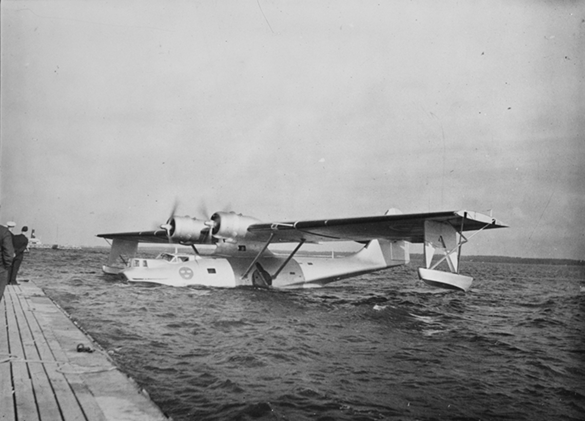Sjöflygplan TP 47 ligger på vatten vid brygga. Till vänster syns personer på bryggan samt ett fartyg i bakgrunden.
