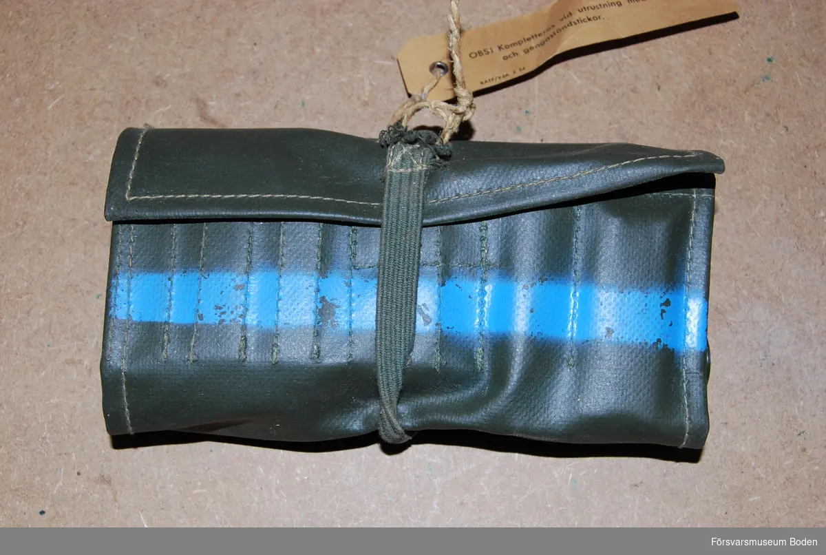 Fodral av grön gummibelagd väv innehållande tändmedelsutrustning (ej helt komplett). Fodralet är märkt med blått band för övningsammunition.