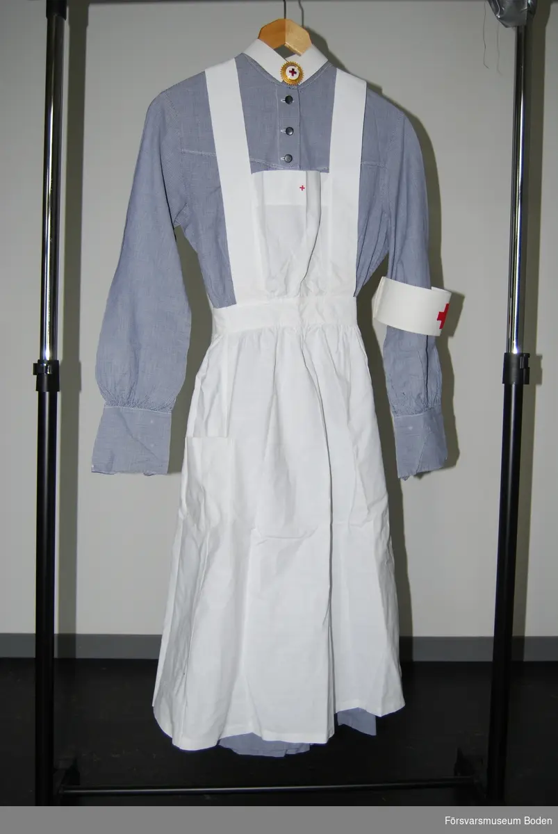 Smårutigt bomullstyg i blått och vitt. Klänningen användes tillsammans med vitt förkläde, mörkblått skärp, vit krage, brosch, armbindel samt på huvudet vit hätta, vilka utgjorde arbetskläderna för en sjuksköterska vid Röda Korset.