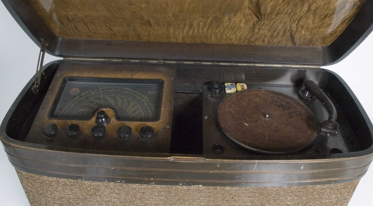 Radiokabinett i tre og trukket med brunmelert stoff. Kabinettet inneholder radio og platespiller.