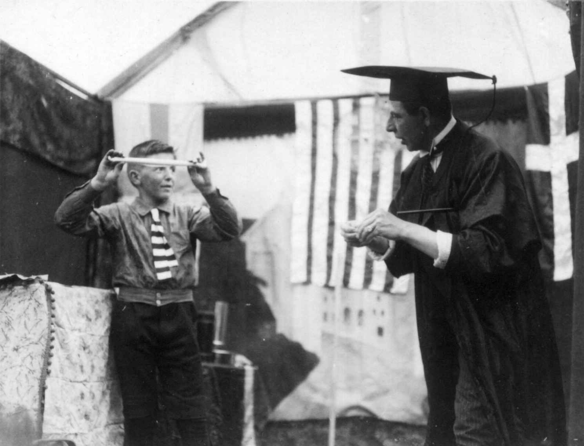 Mann med svart drakt, antakelig tryllekunstner, og en ung gutt. Underholdning på marked på Norsk Folkemuseum i 1924.