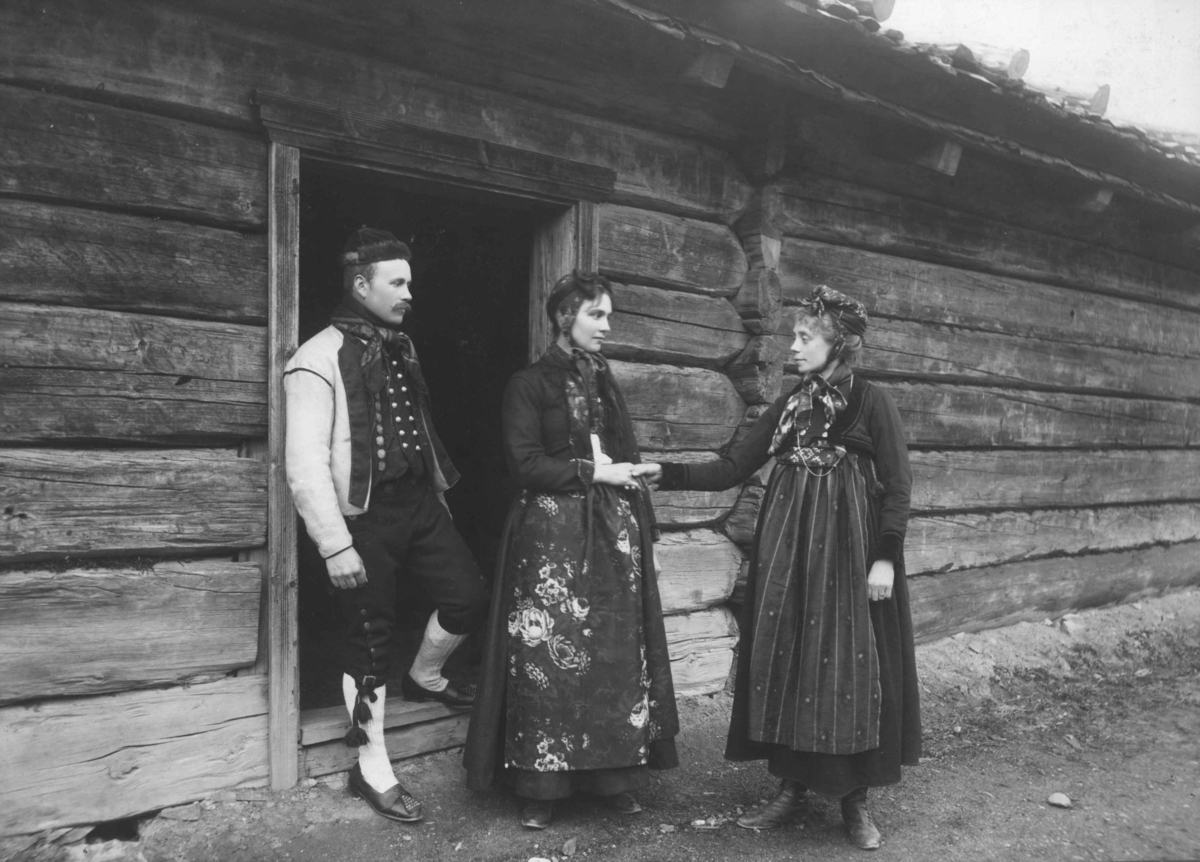 Tilstelning på Norsk Folkemuseum.
En mann og to kvinner i drakter foran et laftet hus på Norsk Folkemuseum