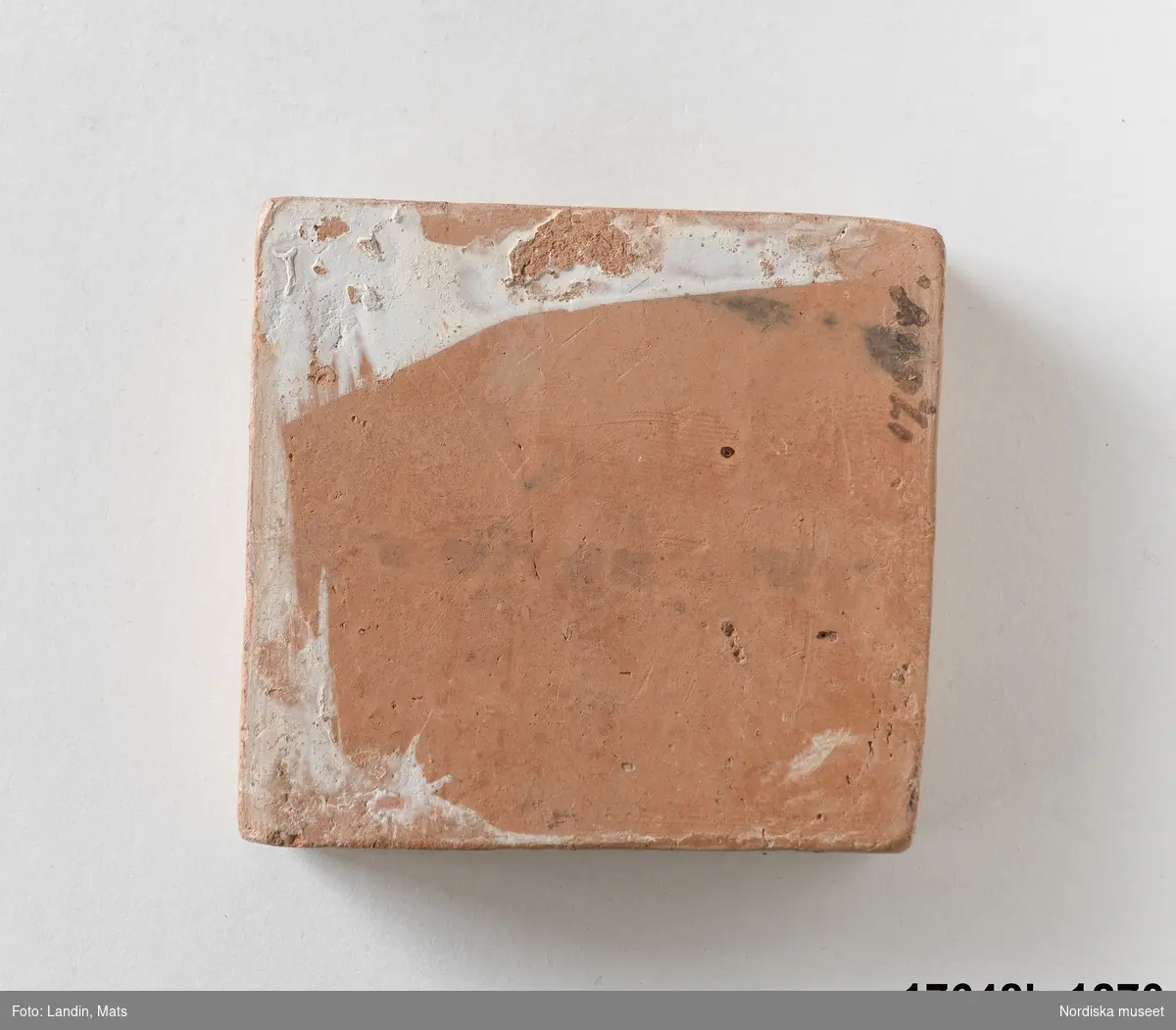 Kvadratisk platta av bränd röd lera, ena sidan glaserad i brungrön (melerat). den andra sidan oglaserad. På den oglaserade sidan text (svagt och därför svårtydd): "Antimon"[?]
/Leif Wallin 2014-01-07