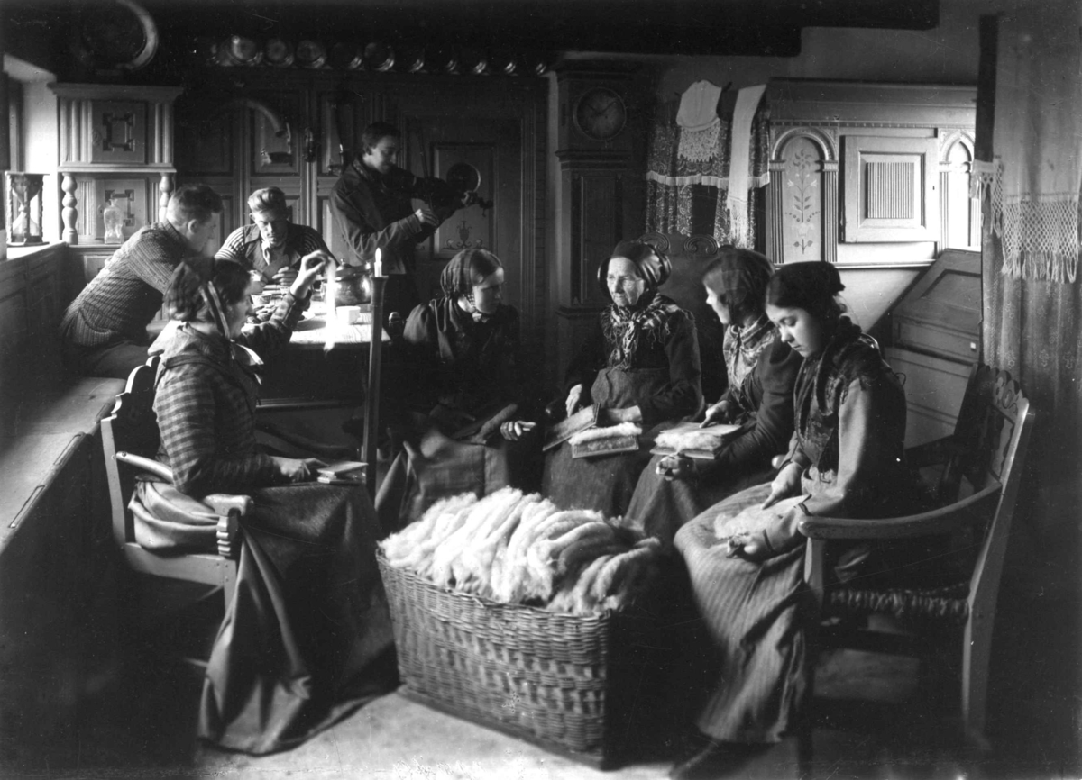 Karding, "kartegilde". Kvinner og menn samlet i en stue og karder ull, en ullkurv i forgrunnen. Stensrup, Danmark.