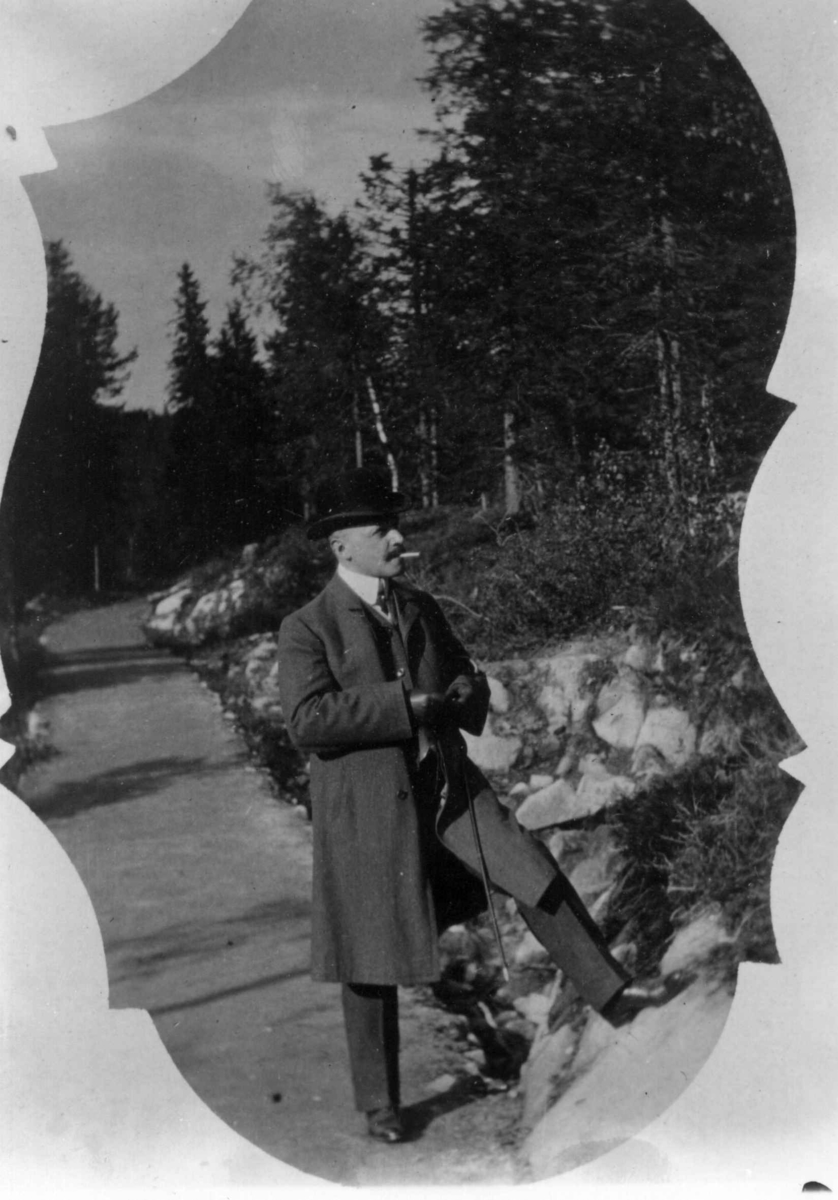 Drakt. Oslo 1908-10. Skog. Vei. I forgrunnen mann i frakk og med hatt. 