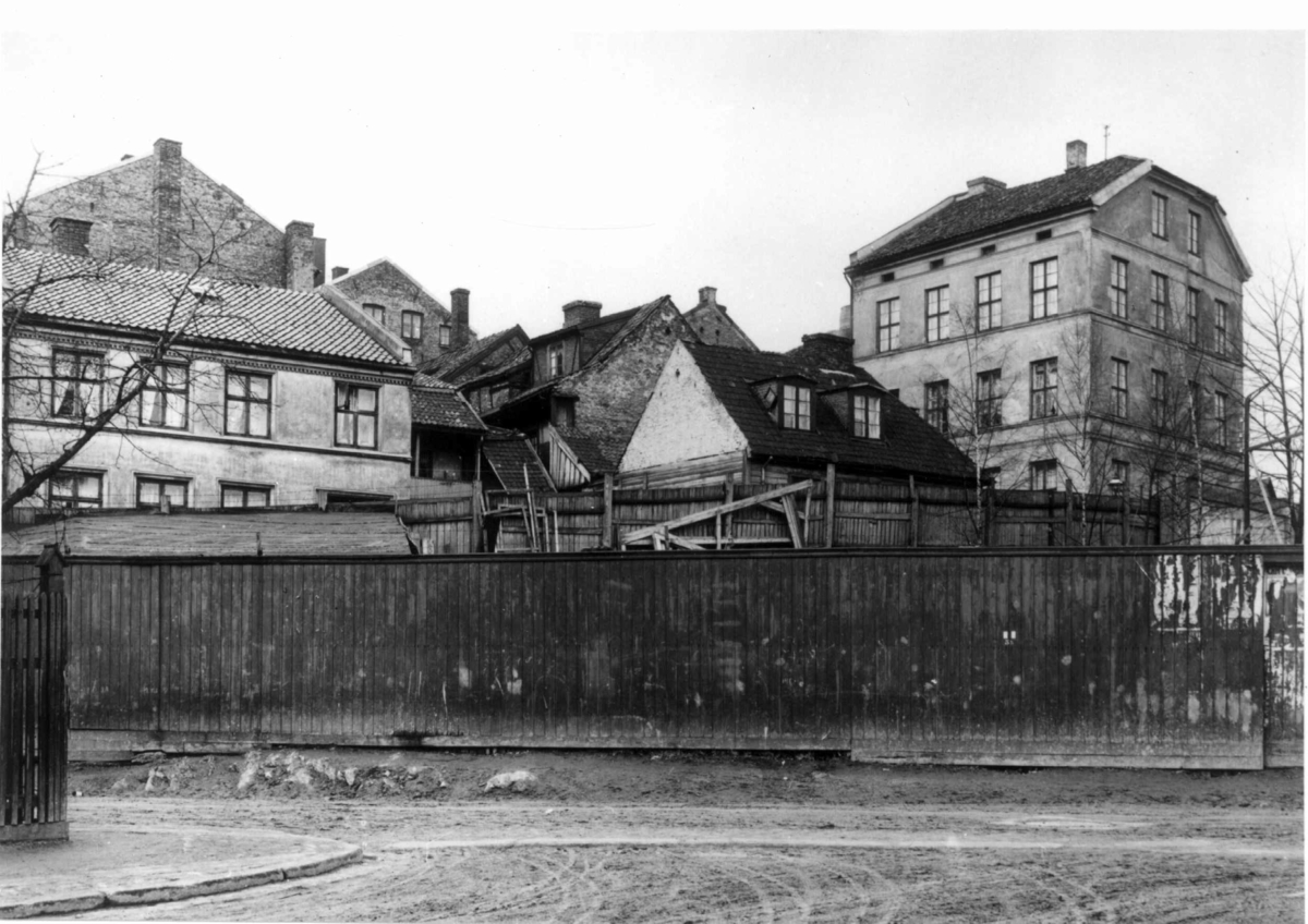 Filosofgangen, Oslo. Gatebilde med bygårder bakenfor plankegjerde. Vinkelgaten i bakgrunnen. Tidligere gate i gamle Vika, før 1866 kalt Pipervigsveien. (Oslo byleksikon.)