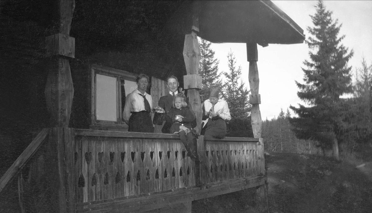 To kvinner, en mann og et barn står på en veranda utenfor en hytte. Robsahm og Lund.