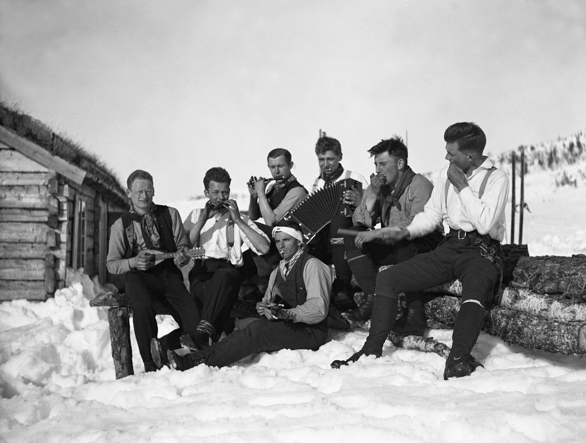 "Høyfjellsorkestret" med syv unge menn spiller opp i snøen utenfor en hytte. Fritjof Arentz med forbinding rundt hodet sitter forrest. Fotografert påsken 1923.