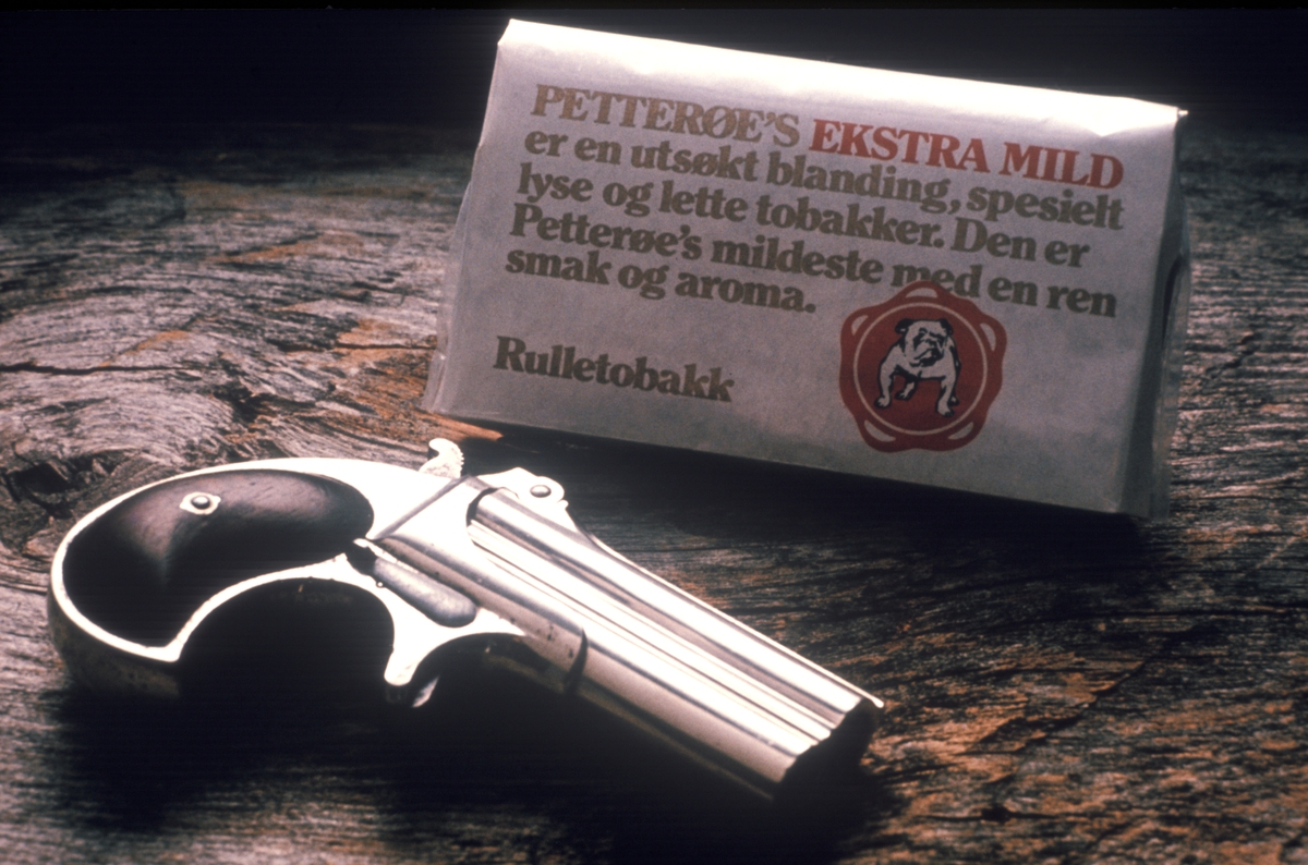 Reklamefoto av Petterøes ekstra mild sammen med pistol. Kampanje for tobakksmerket Petterøes Veteran 1975.