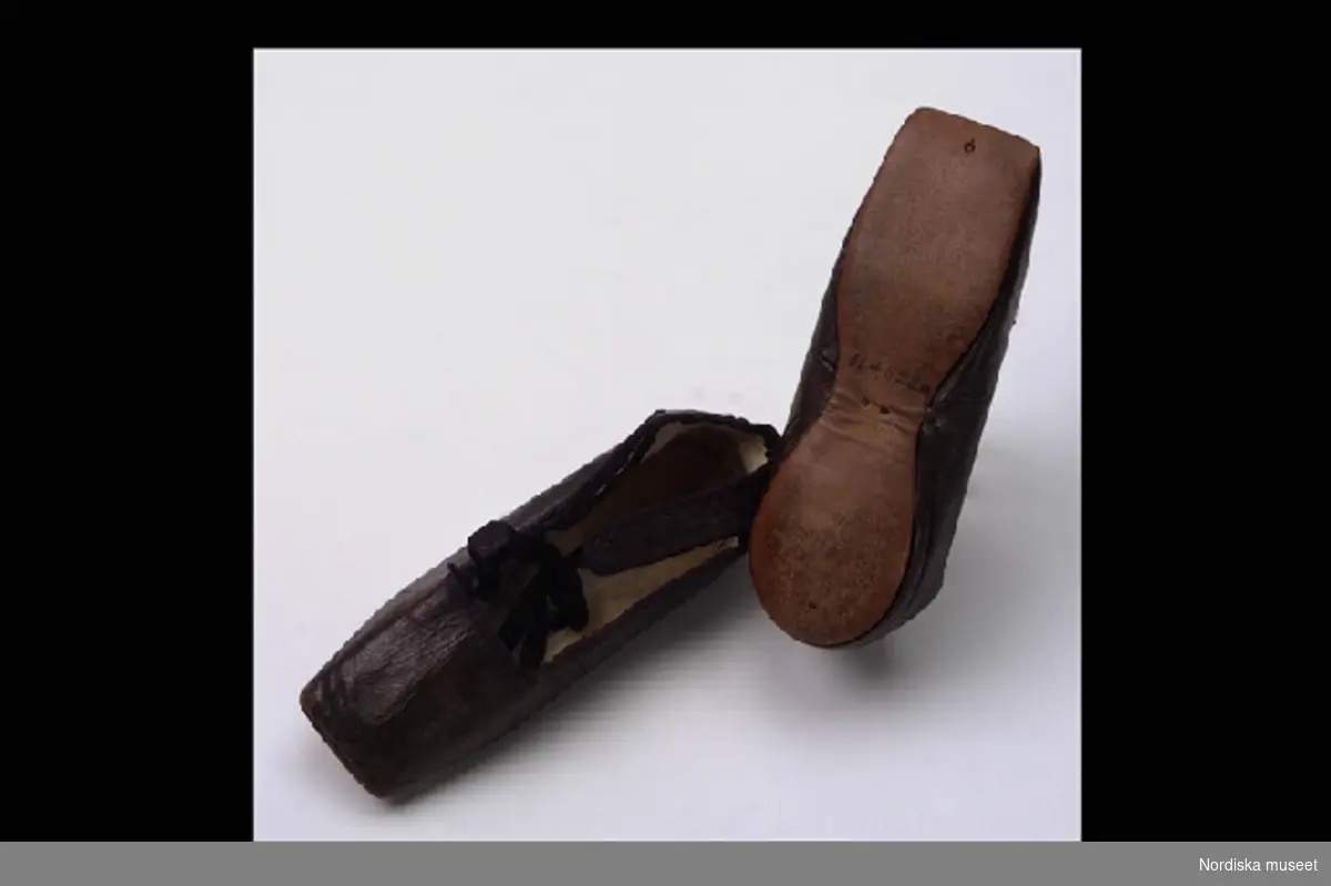 Inventering Sesam 1996-1999:
L  12,5  cm
Skor, dockskor, ett par, ballerinamodell, brunt skinn med platt sula av läder, knytband av svart siden. 
Birgitta Martinius 1996