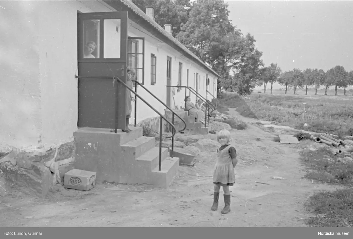 Statarlänga, Bälteberga gård, Skåne. En liten flicka på gårdsplanen framför vitrappad huslänga, andra barn tittar ut ur huset. 