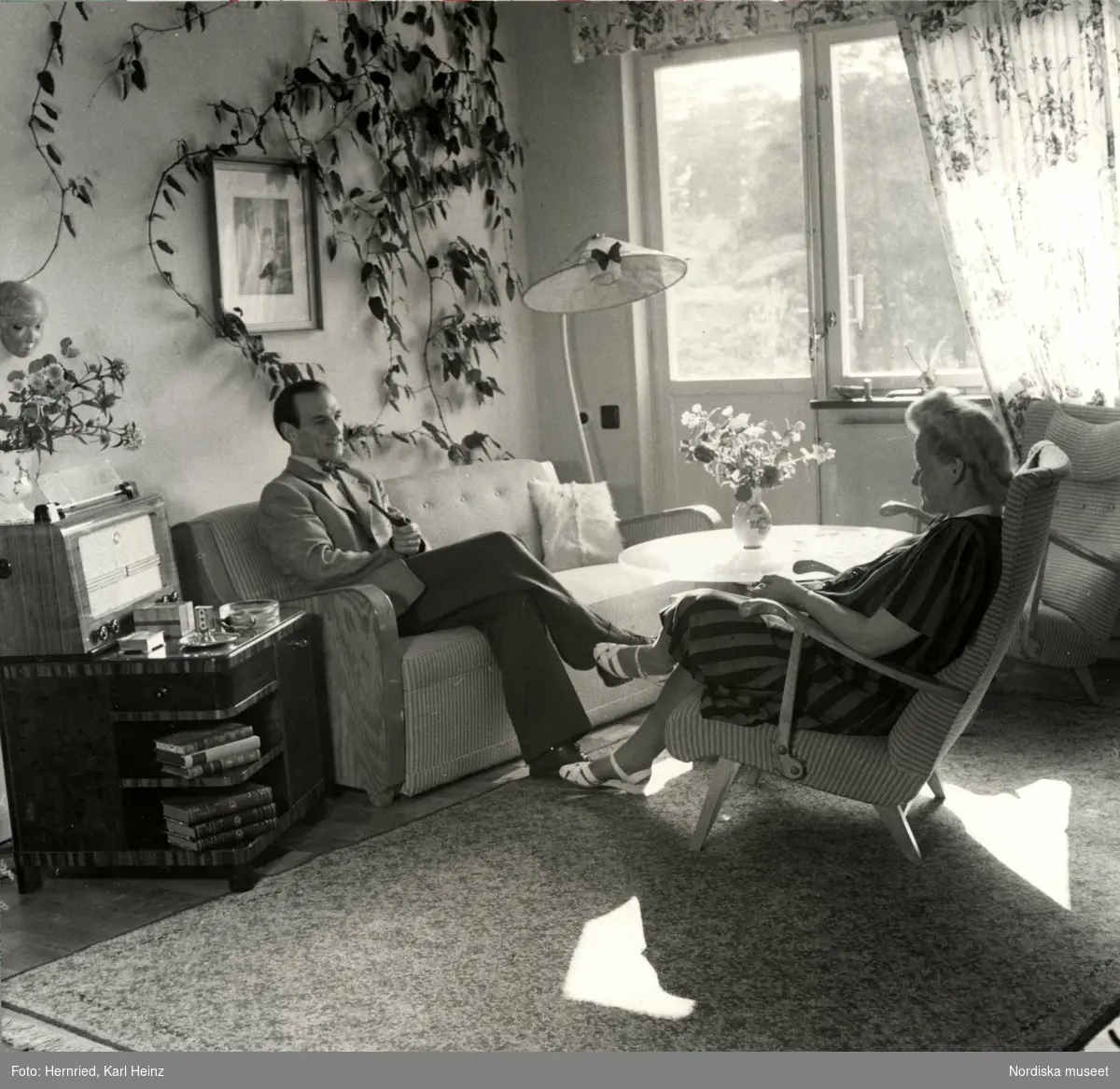 Interiör. Vardagsrum där en kvinna och en man sitter och samtalar, i soffa respektive fåtölj.
