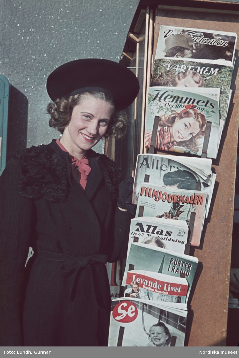 Leende kvinna i svart kappa med pälskrage och hatt bredvid tidningsställ med veckotidningar.